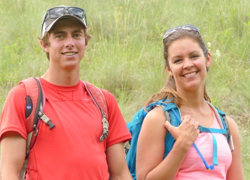 male and female hiking guides stand with backpacks in front of a flowery hillside