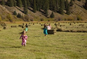 four little girls run toward camera among colorado sunlit hay bales with rocky mountain pines in the distance