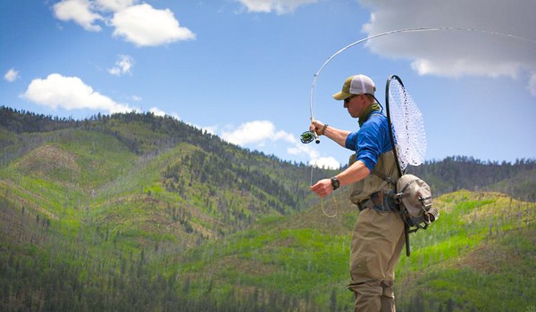 a sunny blue sky with green mountains in the background and a fly fishing guide in the foreground practicing his cast