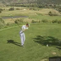 A golfer tees off from an elevated tee box, overlooking a long par 5 hole