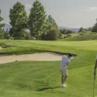 A golfer hits a chip shot towards the green, over a bunker